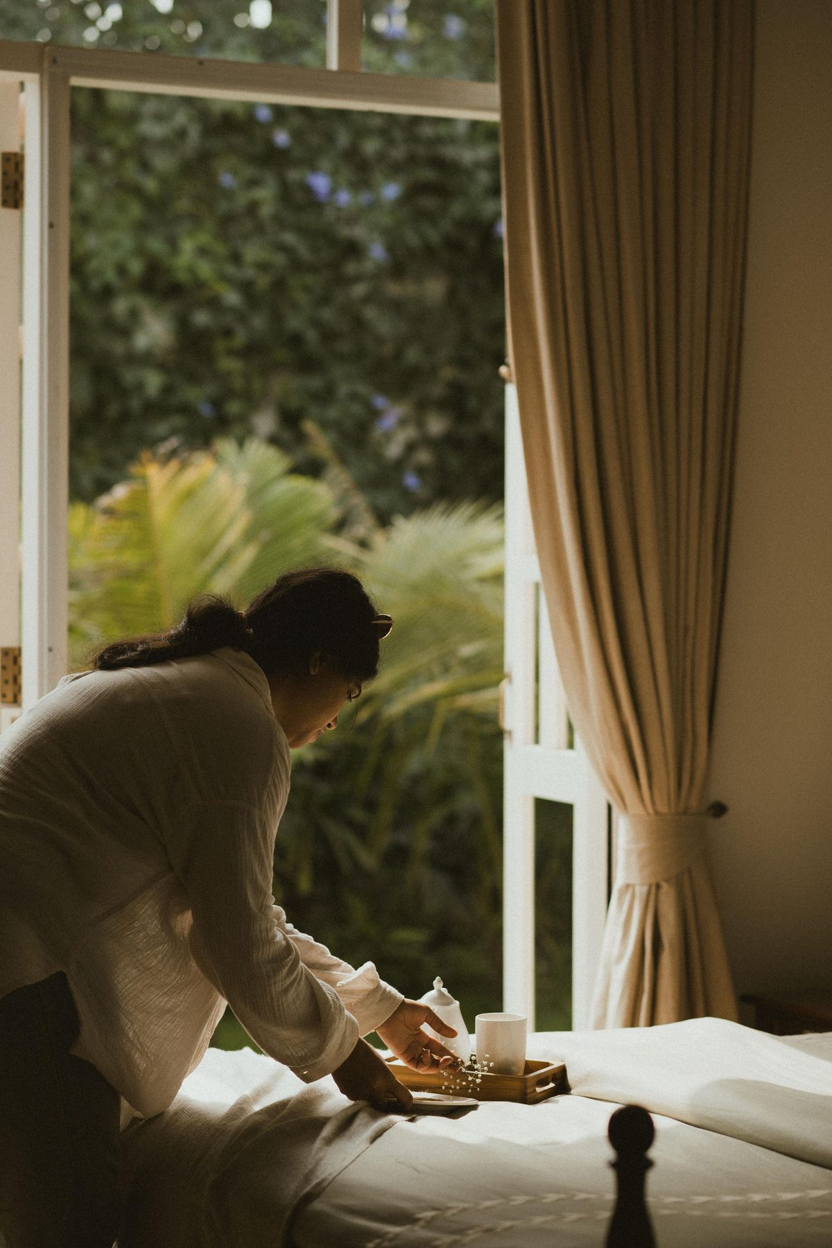 Simran Siriskar arranging a tea tray by an open window, palms and greenery beyond